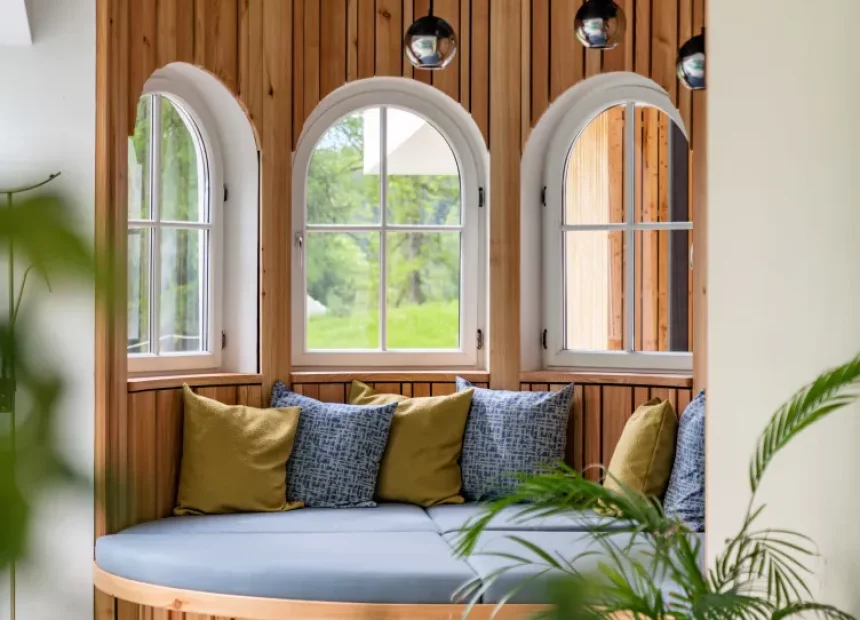 Eine Relaxliege mit gemütlichen Polstern in gedeckten Farben, Holzdetails und einem weißen Erkerventfenster, das den Blick ins Grüne freigibt.