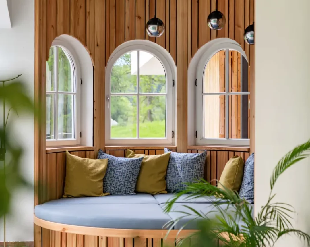Eine Relaxliege mit gemütlichen Polstern in gedeckten Farben, Holzdetails und einem weißen Erkerventfenster, das den Blick ins Grüne freigibt.
