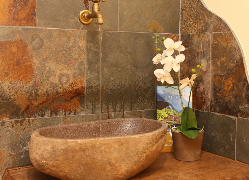 Vor Fliesen aus Naturstein steht ein Waschbecken aus Naturstein mit einem goldenen Wasserhahn. Daneben steht eine weiße Orchidee.