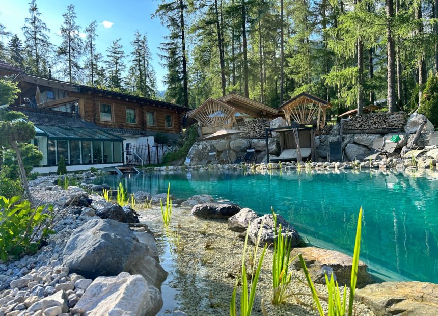 Ein blau-grüner Naturteich, umgeben von Steinen und grünen Bäumen. Im Hintergrund ist das Naturhotel Lärchenhof in Ramsau am Dachstein zu sehen.